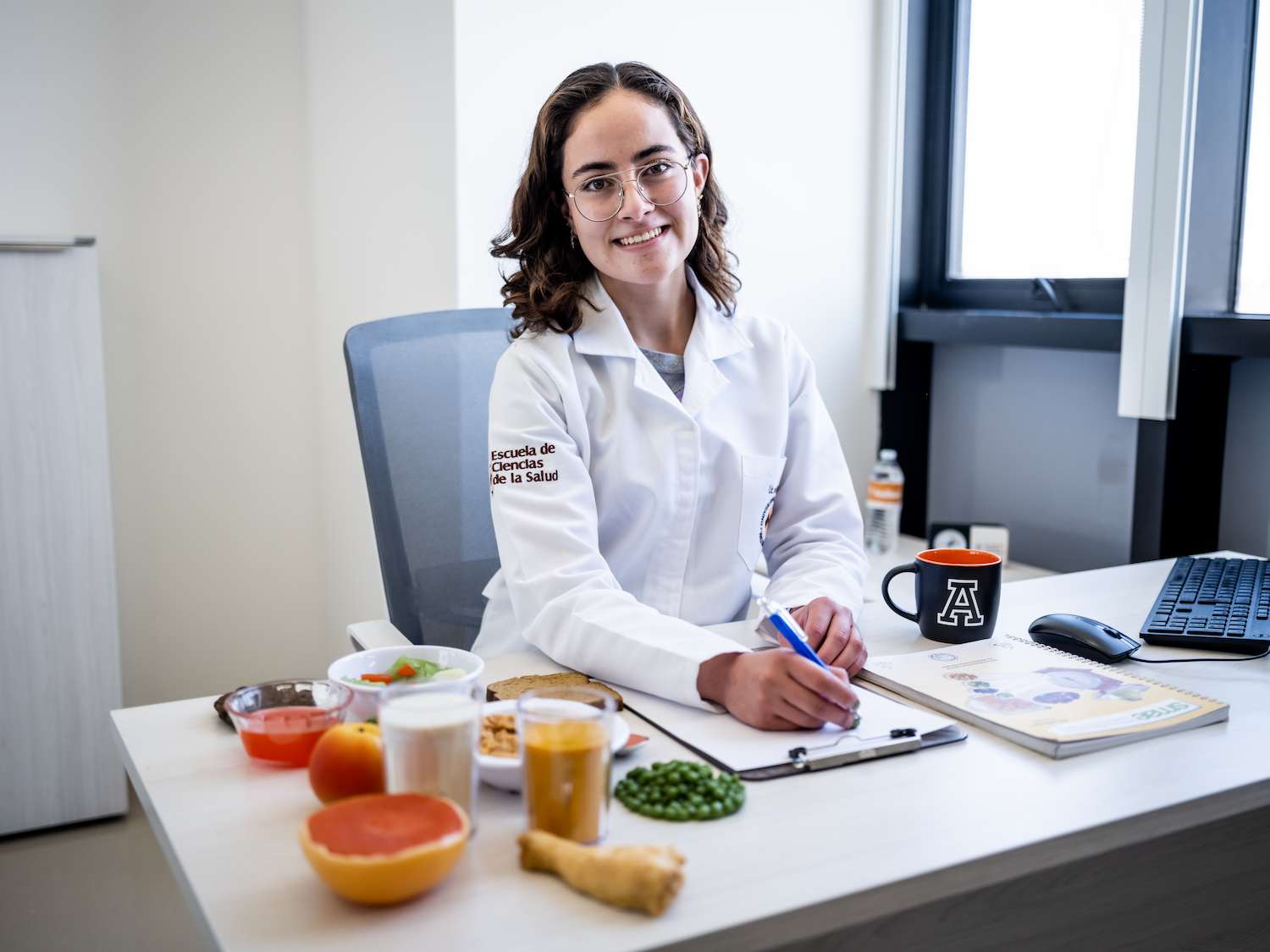 Nutrióloga sonriente toma notas en escritorio con alimentos variados, libros de nutrición y taza institucional.